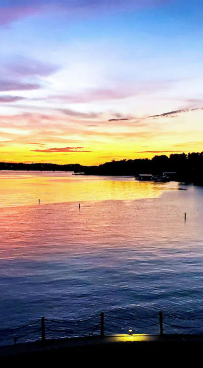 Vibrant sunset over a calm lake, orange, pink and blue sky reflected on the water with silhouetted tree-lined shoreline and small docks in the distance.