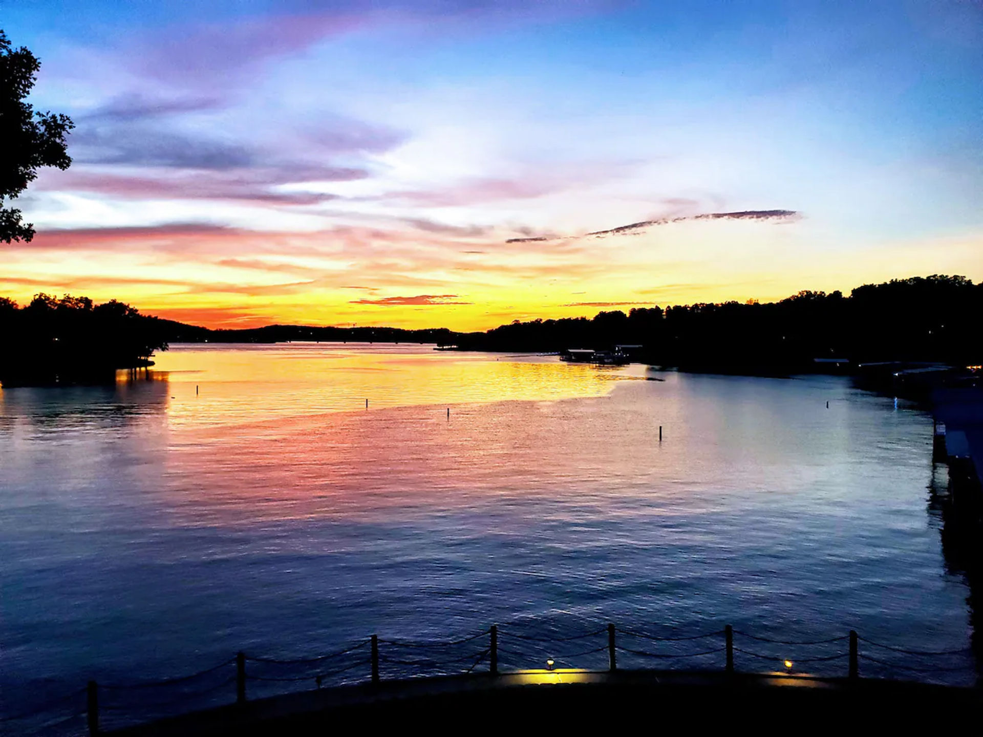 Vibrant sunset over a calm lake, orange, pink and blue sky reflected on the water with silhouetted tree-lined shoreline and small docks in the distance.