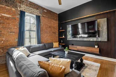 Cozy industrial loft living room with exposed brick wall, arched window with navy curtains, large gray sectional, leather accent pillows, dark wood coffee table, and wall-mounted TV on a modern black-and-wood media wall.