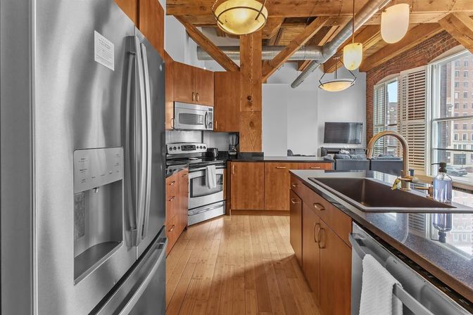 Modern downtown loft kitchen with exposed wooden beams and ductwork, stainless steel appliances, warm wood cabinets, center island with sink, pendant lights and large city-view windows.