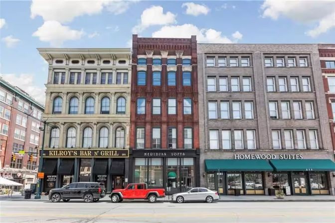 Sunlit historic downtown commercial buildings with ornate stone and red brick facades, street-level storefronts and awnings, parked cars along a city street under a blue sky