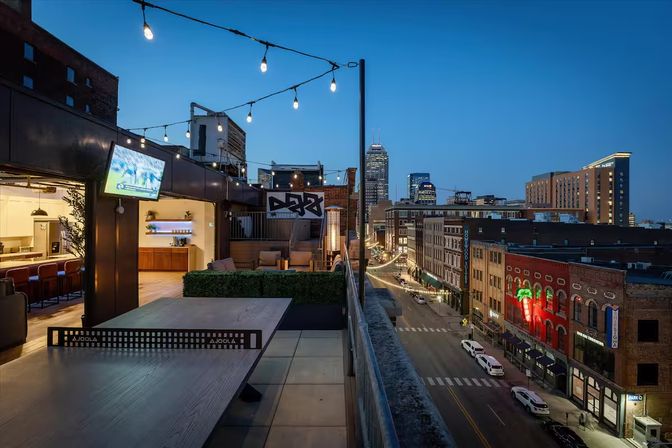 Rooftop terrace at dusk with string lights, outdoor ping-pong table and cozy lounge seating, overlooking a lively downtown street with neon signs and skyline towers.