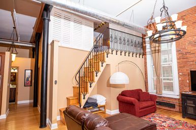 Cozy downtown loft living room with exposed brick wall and tall windows, wrought-iron staircase to a mezzanine, leather sofa and red armchair on hardwood floors, industrial ductwork and chandelier