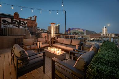 Cozy urban rooftop lounge at dusk with cushioned sofas and armchairs around a modern fire pit and tall patio heater, string lights overhead and a lit downtown skyline and arena in the background.