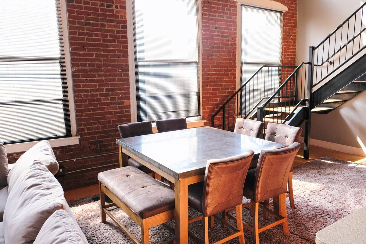 Sunlit urban loft dining area with exposed red brick walls, tall windows with blinds, a square metal-top dining table surrounded by leather chairs and a tufted bench, and a black metal staircase.
