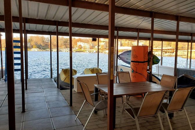 Covered lakeside dock with a wooden dining table and several chairs, stacked paddleboards and two yellow personal watercraft in a covered boat slip, overlooking a calm lake and shoreline homes in warm late-afternoon light.