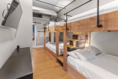 Loft-style shared bedroom with double wooden bunk beds, crisp white linens, built-in wooden steps, exposed industrial ductwork, wall-mounted TV and light hardwood floors