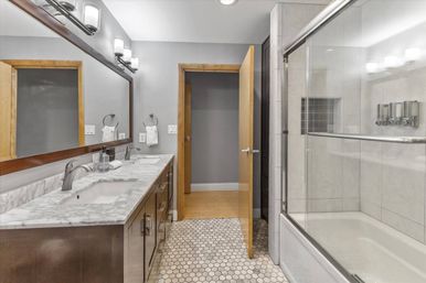 Sleek modern bathroom with marble double vanity and undermount sinks, large wood-framed mirror, hexagon tile floor, glass-enclosed tub/shower, gray walls, wood door and chrome fixtures.