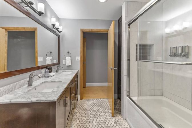 Sleek modern bathroom with marble double vanity and undermount sinks, large wood-framed mirror, hexagon tile floor, glass-enclosed tub/shower, gray walls, wood door and chrome fixtures.