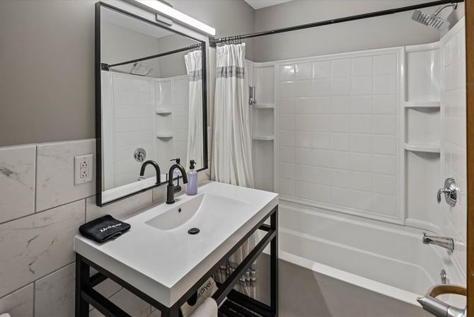 Sleek modern bathroom with white tub-shower combo and tiled surround, black-framed mirror, matte-black sink faucet on a white vanity, striped shower curtain and neutral gray walls.