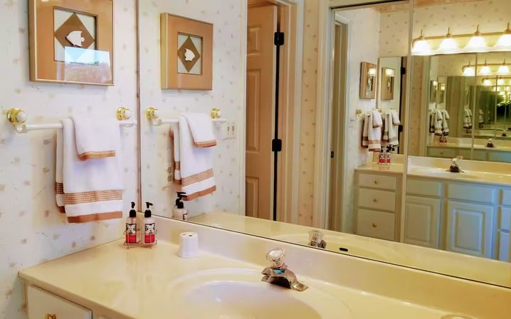 Bright residential bathroom vanity with cream sink and countertop, large wall mirror reflecting brass lights, neatly folded white towels with tan trim and soap dispensers on the counter.