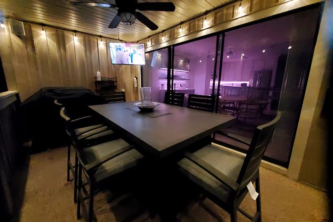 Cozy covered backyard patio at night with a modern high-top dining table and cushioned bar stools, string lights and ceiling fan, wall-mounted TV, and sliding glass doors revealing a purple-lit kitchen inside.