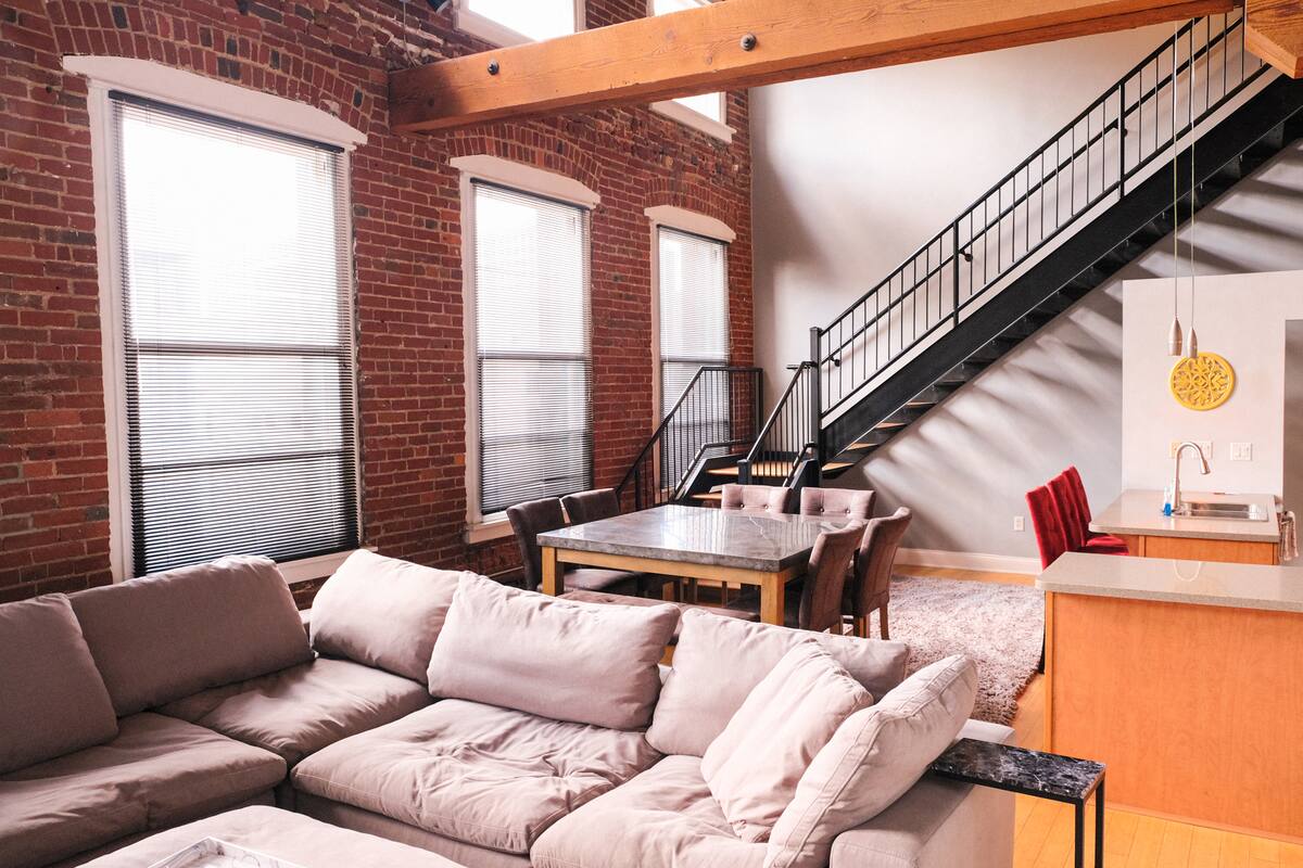 Open-plan urban loft with exposed red brick wall and tall windows, a cozy beige sectional, wooden beam, dining table, black metal staircase, and a compact kitchen island with red barstools.