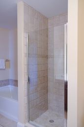 Corner beige tiled walk-in shower with frameless glass door and chrome handle next to a white bathtub in a modern residential bathroom.