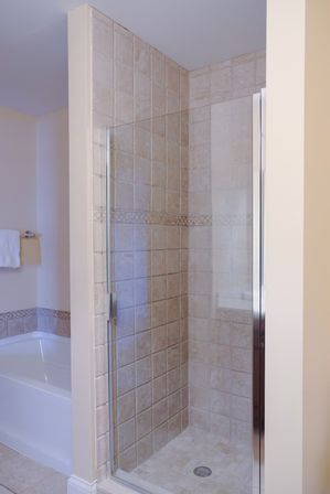 Corner beige tiled walk-in shower with frameless glass door and chrome handle next to a white bathtub in a modern residential bathroom.