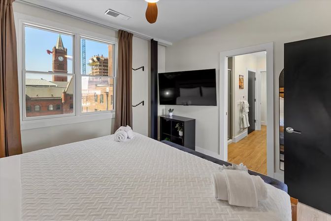Sunny modern downtown bedroom with white quilted bed and rolled towels, large window framing brick buildings and a clock tower, wall-mounted TV and open doorway to en-suite bathroom.