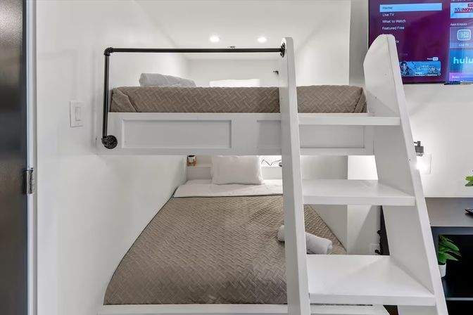 Compact modern white bunk beds with ladder and gray quilted bedding in a small apartment bedroom with wall-mounted TV