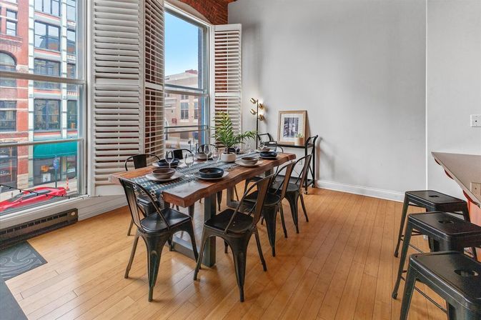 Bright downtown loft dining area with a rustic wooden table set for six, black metal chairs, hardwood floors and large bay windows overlooking a city street.