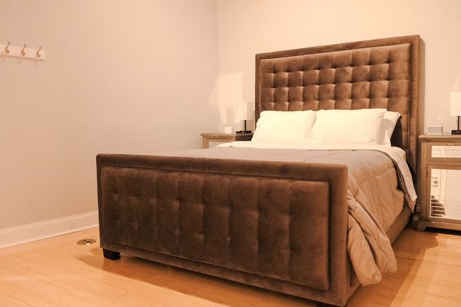 Cozy modern bedroom interior with a tufted brown upholstered bed, white pillows and beige comforter, mirrored nightstands and lamps on light hardwood floor.