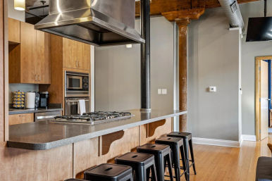 Modern industrial-style loft kitchen with a long gray countertop island, built-in gas cooktop under a stainless steel hood, wooden cabinets, black metal bar stools, exposed ductwork and hardwood floors.