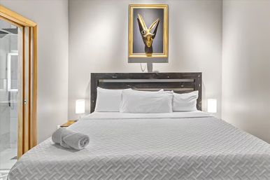 Minimalist modern bedroom with a king bed dressed in white quilted linens and pillows, rustic dark wood headboard, rolled towel on the bed, framed gold antelope skull wall art, bedside lamps, and a glimpse of a glass-walled ensuite bathroom.