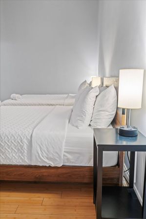 Crisp modern guest room with two twin beds dressed in white linens, wooden platform frame, black nightstand and a glowing bedside lamp against light gray walls and hardwood floor