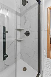 Sleek glass-enclosed shower with white marble-effect tile walls, matte-black rain showerhead and control, two corner shelves, white shower base, and adjacent hexagon-tile bathroom floor with wooden doorframe.