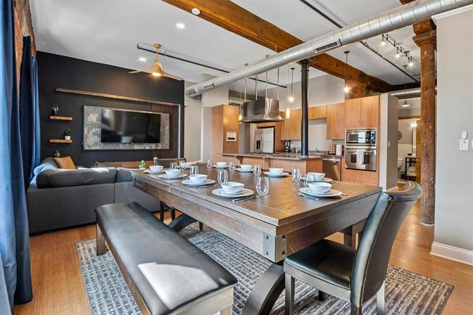 Industrial urban loft dining area with a large wooden farmhouse table set for eight, open-concept kitchen with stainless steel appliances, exposed wooden beams and ductwork, and a cozy gray sectional living area.