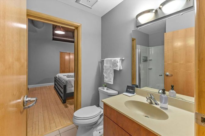 Clean modern en-suite bathroom with gray walls and wood-trimmed door opening to a bedroom with hardwood floors, single-sink vanity with soap dispenser, toilet, towel on wall rack, and glass-enclosed shower.