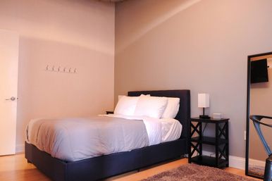 Cozy minimalist apartment bedroom with navy upholstered queen bed, gray duvet and white pillows, black nightstand with lamp, full-length mirror and light wood floors