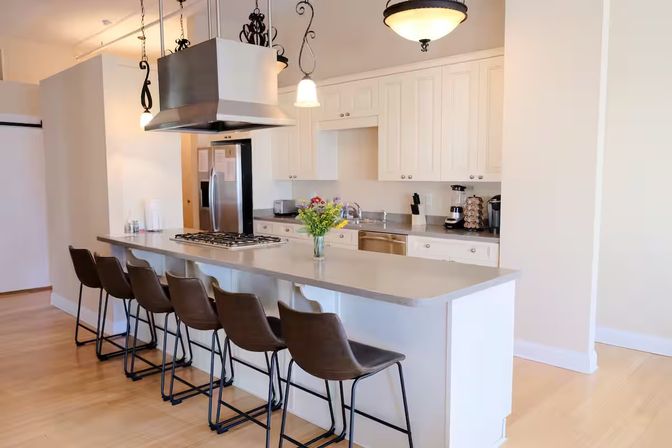 Bright open-concept modern kitchen with long gray island and five brown bar stools, stainless steel range hood and appliances, white cabinetry, pendant lights, hardwood floors and a vase of fresh flowers on the counter.