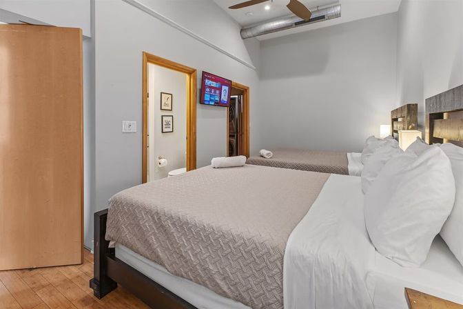 Bright modern urban loft guest room with two beds, neutral bedding, wall-mounted TV, ceiling fan and stacked washer-dryer visible.