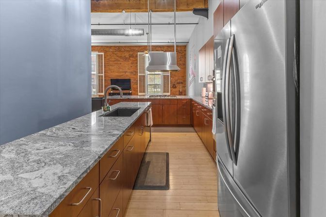 Bright urban loft-style kitchen with exposed brick, stainless-steel French-door refrigerator, gray marble island with sink, warm wood cabinets, pendant range hood and light hardwood floors.