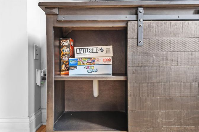 Farmhouse-style wooden console with sliding barn door open to reveal stacked board games (Battleship, Jenga, Life) on a living-room shelf, family game-night ready.