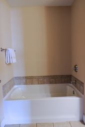 White alcove bathtub with beige tile trim and mosaic border, chrome faucet and towel bar holding folded white towels against neutral beige bathroom walls and tiled floor