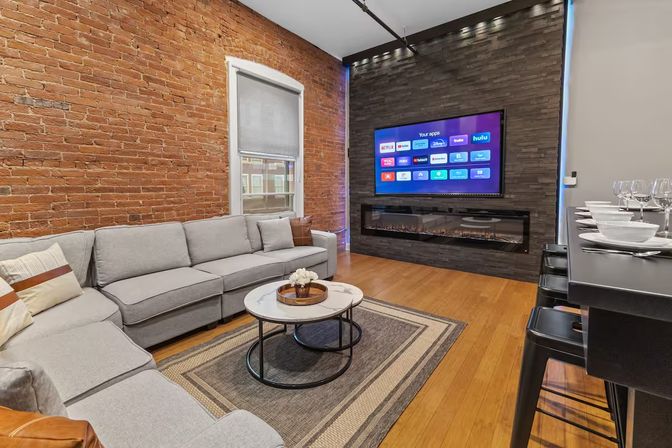Loft-style living room with exposed brick wall, gray sectional sofa, round marble coffee table, wood floors, wall-mounted TV and linear electric fireplace, plus a breakfast bar with stools.