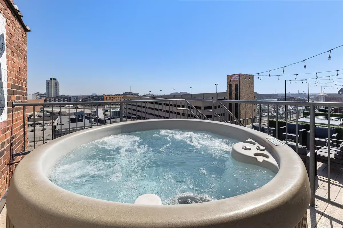 Bubbling rooftop hot tub with railing and string lights, overlooking an urban downtown skyline under a clear blue sky.