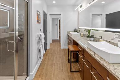 Contemporary spa-like bathroom with double white vessel sinks on a granite countertop, large illuminated mirror, wood-look tile floor, and glass-enclosed walk-in shower.