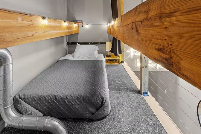 Cozy industrial loft sleeping nook on a mezzanine with a single bed in a gray quilt, exposed wooden beams, visible metal ductwork and warm string lights