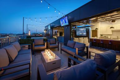 Rooftop lounge at dusk with cozy outdoor seating around a glass fire pit, string lights overhead, wall-mounted TVs and an open bar/kitchen overlooking an urban skyline.