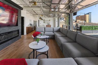 Cozy sunlit urban rooftop lounge with a long gray sectional sofa, nested round marble-top coffee tables with potted plants, wood floors, wall-mounted TV above a linear fireplace, and open glass garage-style doors opening to a city skyline terrace.