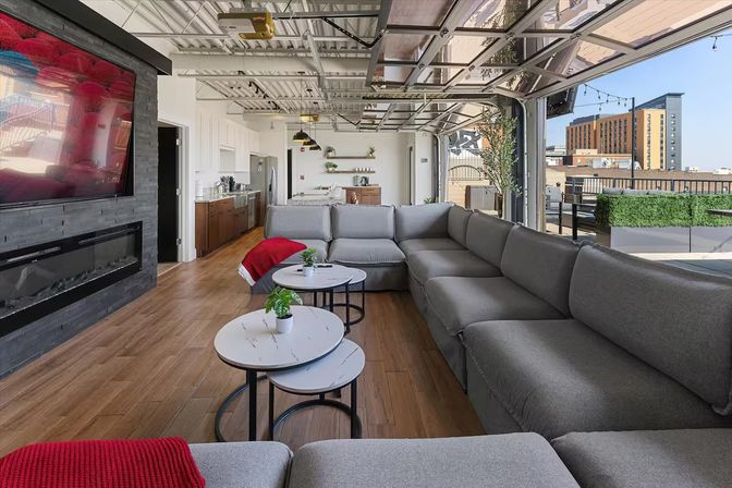 Cozy sunlit urban rooftop lounge with a long gray sectional sofa, nested round marble-top coffee tables with potted plants, wood floors, wall-mounted TV above a linear fireplace, and open glass garage-style doors opening to a city skyline terrace.