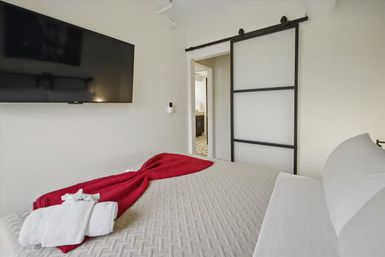 Modern bedroom with textured gray bedspread, cozy red throw and folded white towels on the bed, large wall-mounted flat-screen TV, and black-framed sliding barn door opening to an en-suite bathroom