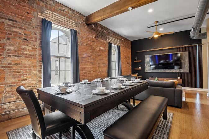 Industrial loft interior with exposed brick walls and arched windows, dark wood dining table set with white dinnerware, bench and leather chairs, exposed ceiling beam and ductwork, hardwood floors and adjacent gray sofa with wall-mounted TV