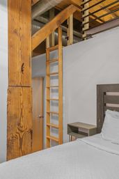 Industrial loft bedroom with vertical wooden ladder to a mezzanine, exposed rustic timber beams and silver ductwork, gray nightstand and white bedding