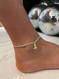 Close-up of a bare ankle wearing a twisted silver anklet with a small gold charm, mirrored disco balls reflecting light in the blurred background.