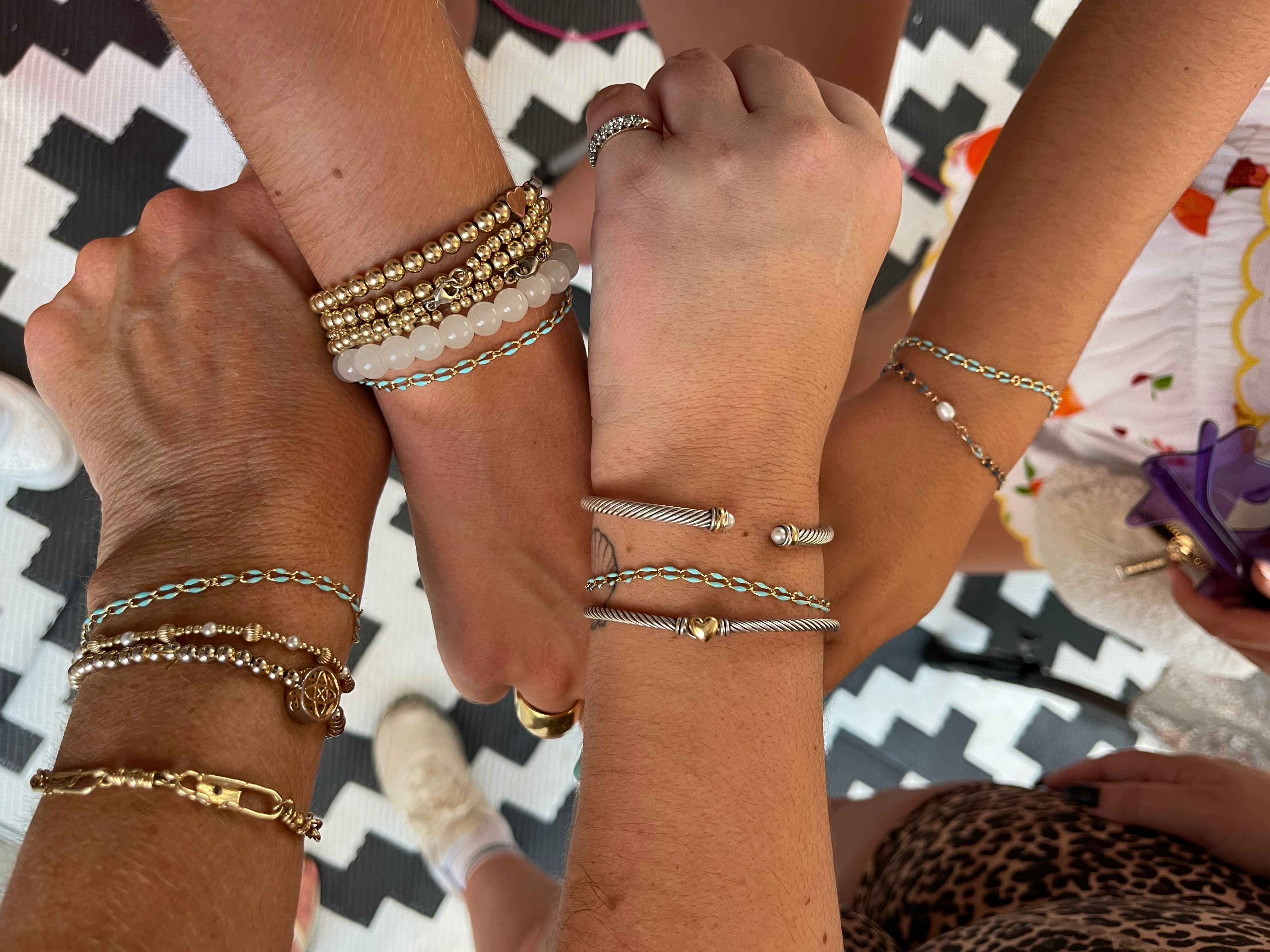 Four wrists together showing stacked bracelets and rings — mixed-metal cable cuffs, gold beads, turquoise-accented chains and white beads on a black-and-white chevron rug.