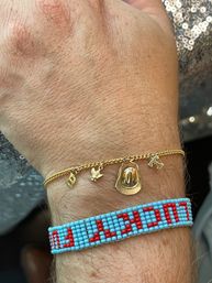 Close-up of a wrist wearing a delicate gold charm bracelet with a cowboy hat, small bird and letter charms paired with a turquoise-and-red beaded friendship bracelet
