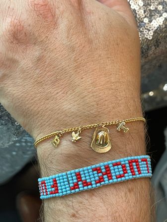 Close-up of a wrist wearing a delicate gold charm bracelet with a cowboy hat, small bird and letter charms paired with a turquoise-and-red beaded friendship bracelet