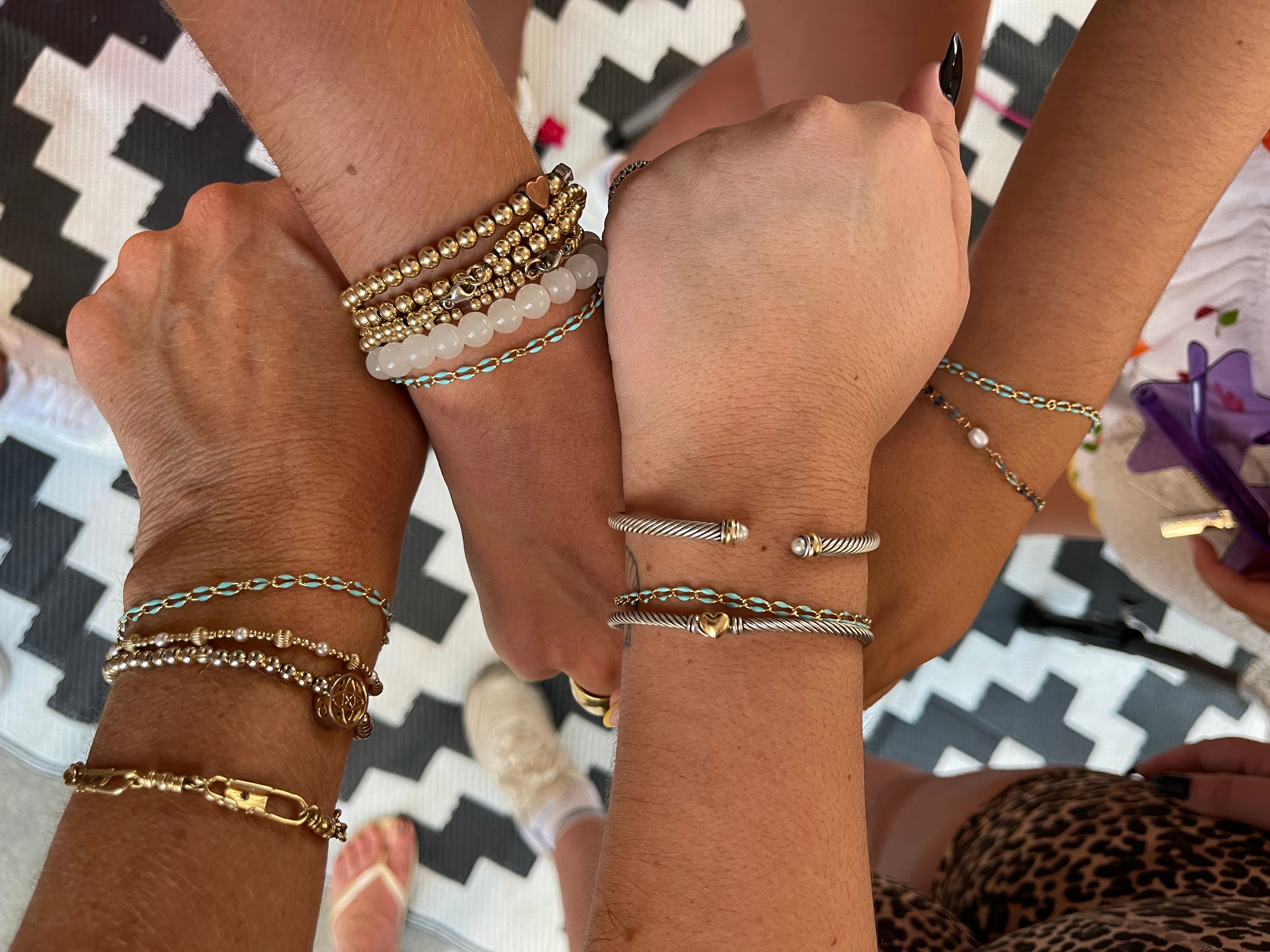 Close-up top-down view of several wrists joined in a circle showing stacked gold, silver and beaded bracelets, twisted cable bangles with a small heart charm, on a black-and-white chevron rug.